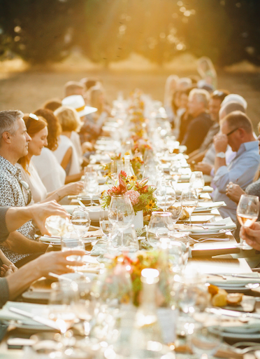 A long table full of people with golden light in the background. The table is full of wine glasses and flowers.